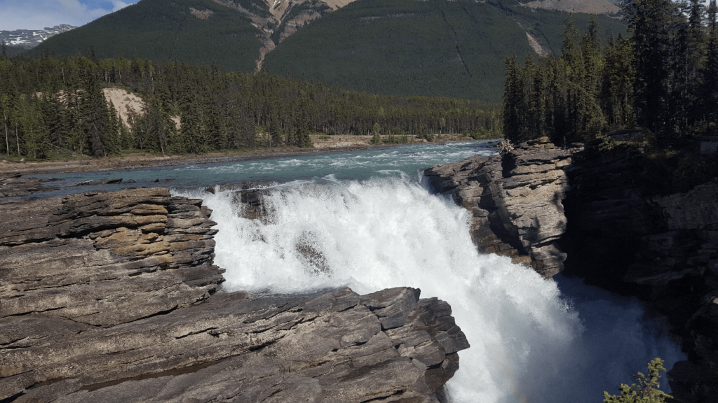large waterfall with craggy rocks on each side, with a forested hill in the background