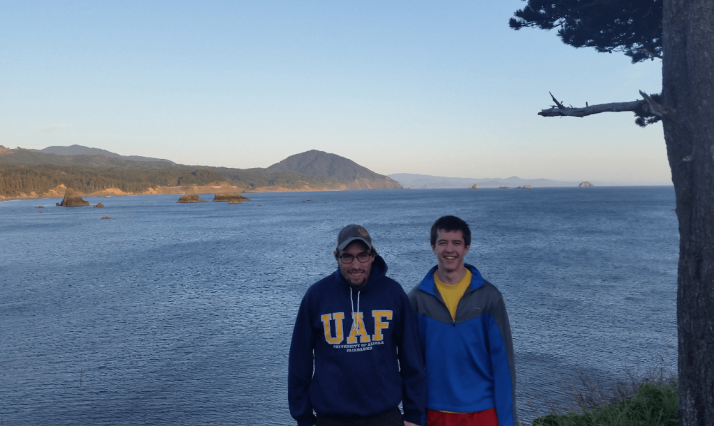 Mike and Joseph next to a pine tree with the oregon coast behind